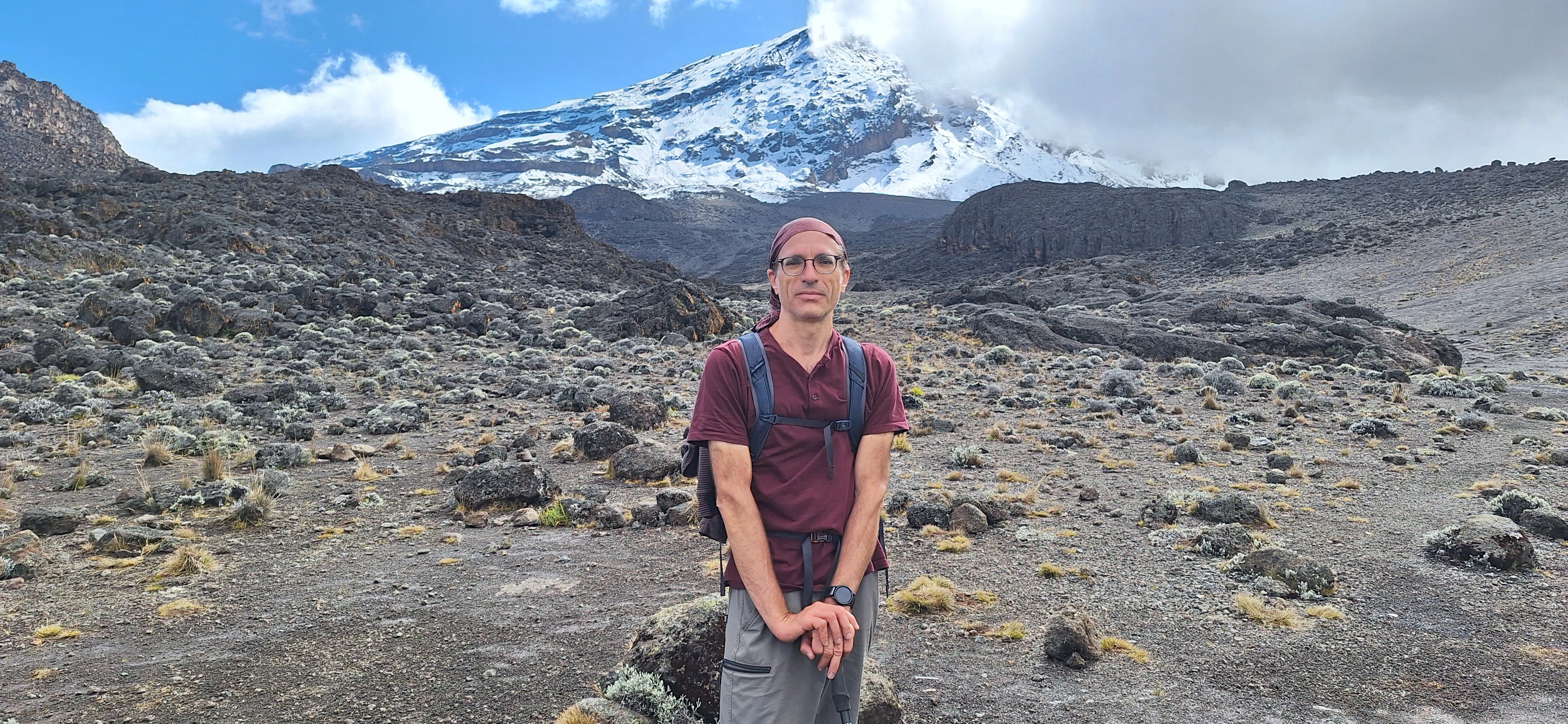 Richard Massel wearing a hiking pack with a snow-capped mountain peak behind him.
