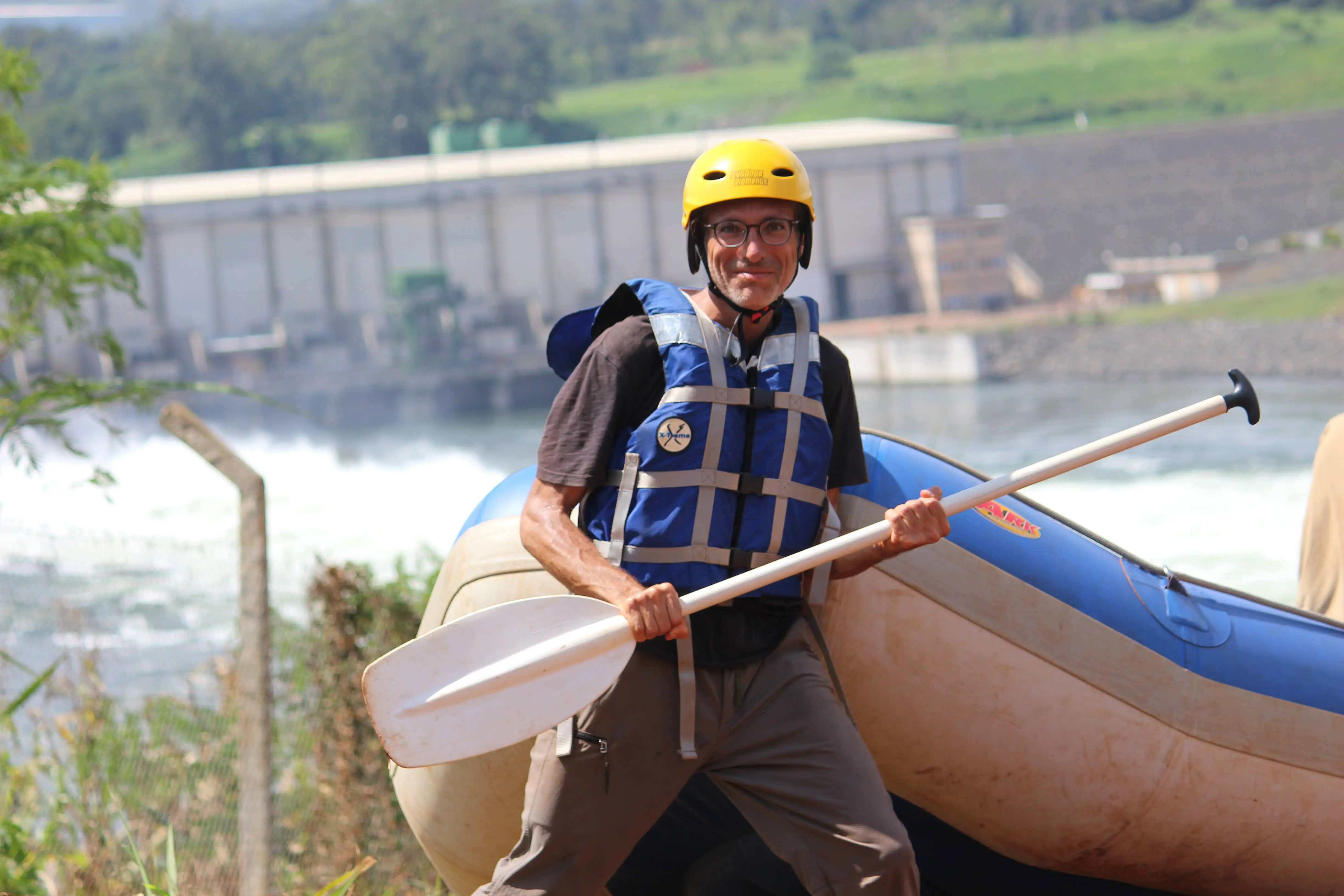 Richard Massel wearing a yellow helmet and holding a paddle next to a whitewater raft near the Source of the Nile.